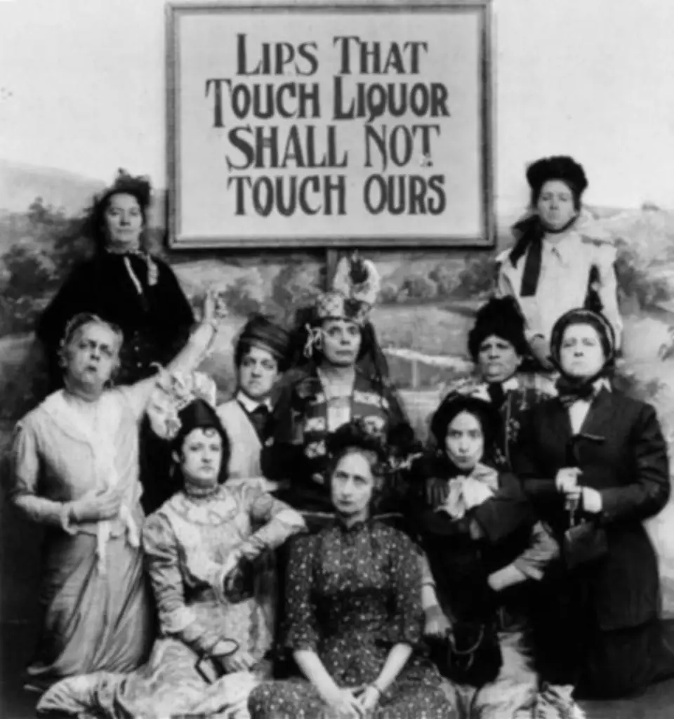 Black and white historical photograph of a group of nine women from the Temperance movement. They are posed in two rows, some seated and some standing, with solemn expressions. Behind them, a large sign reads 'Lips That Touch Liquor Shall Not Touch Ours,' indicating their commitment to the cause against alcohol consumption.