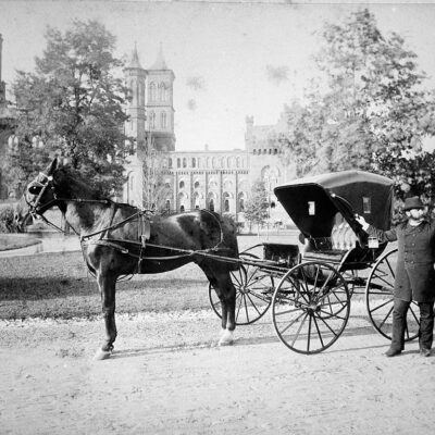 Daniel H. Riggs, Howard University Medicine, in the South Yard of the Smithsonian Institution Building, stands next to his carriage. Picture shows the East Wing after Adolph Cluss' 1884 renovation. Against the building are sheds, used by the Department of Living Animals, 1887-1890 (Smithsonian Archives)