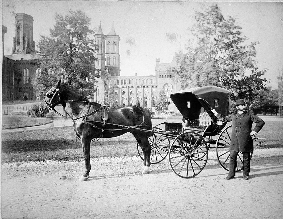 Daniel H. Riggs, Howard University Medicine, in the South Yard of the Smithsonian Institution Building, stands next to his carriage. Picture shows the East Wing after Adolph Cluss' 1884 renovation. Against the building are sheds, used by the Department of Living Animals, 1887-1890 (Smithsonian Archives)