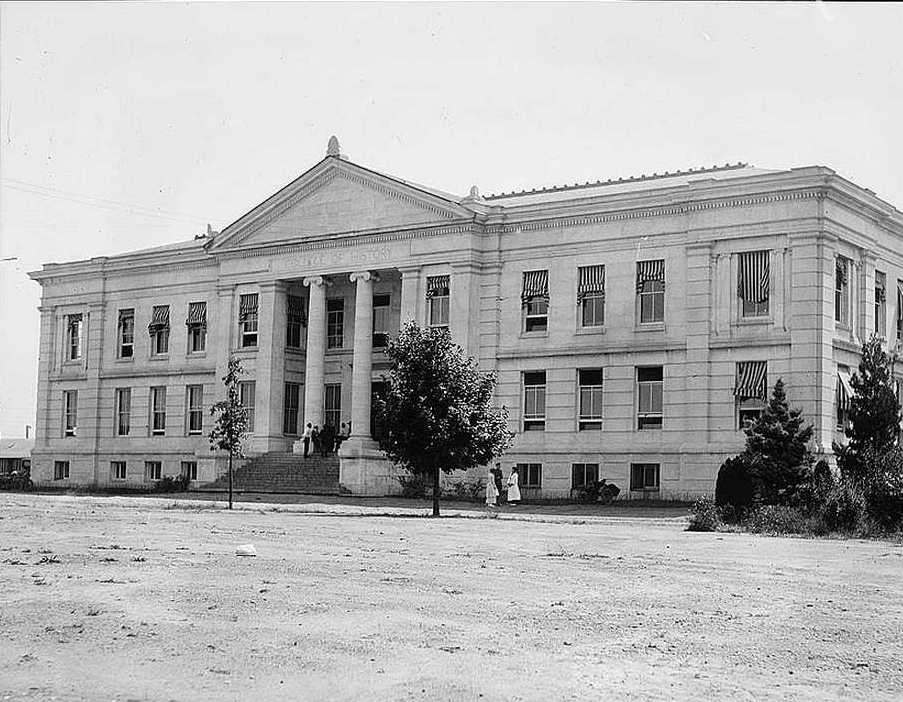 Black and white photograph of Hurst Hall at American University, featuring a neoclassical architectural style with a prominent central pediment supported by large columns, large symmetrical windows, and American flags hanging from the building. A few individuals are visible at the entrance and trees flank the sides.