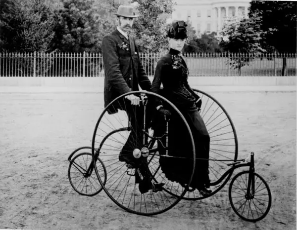 Smartly dressed couple seated on an 1886-model bicycle for two. The South Portico of the White House, Washington, D.C., in the background. 77-RP-7347-4. (National Archives)