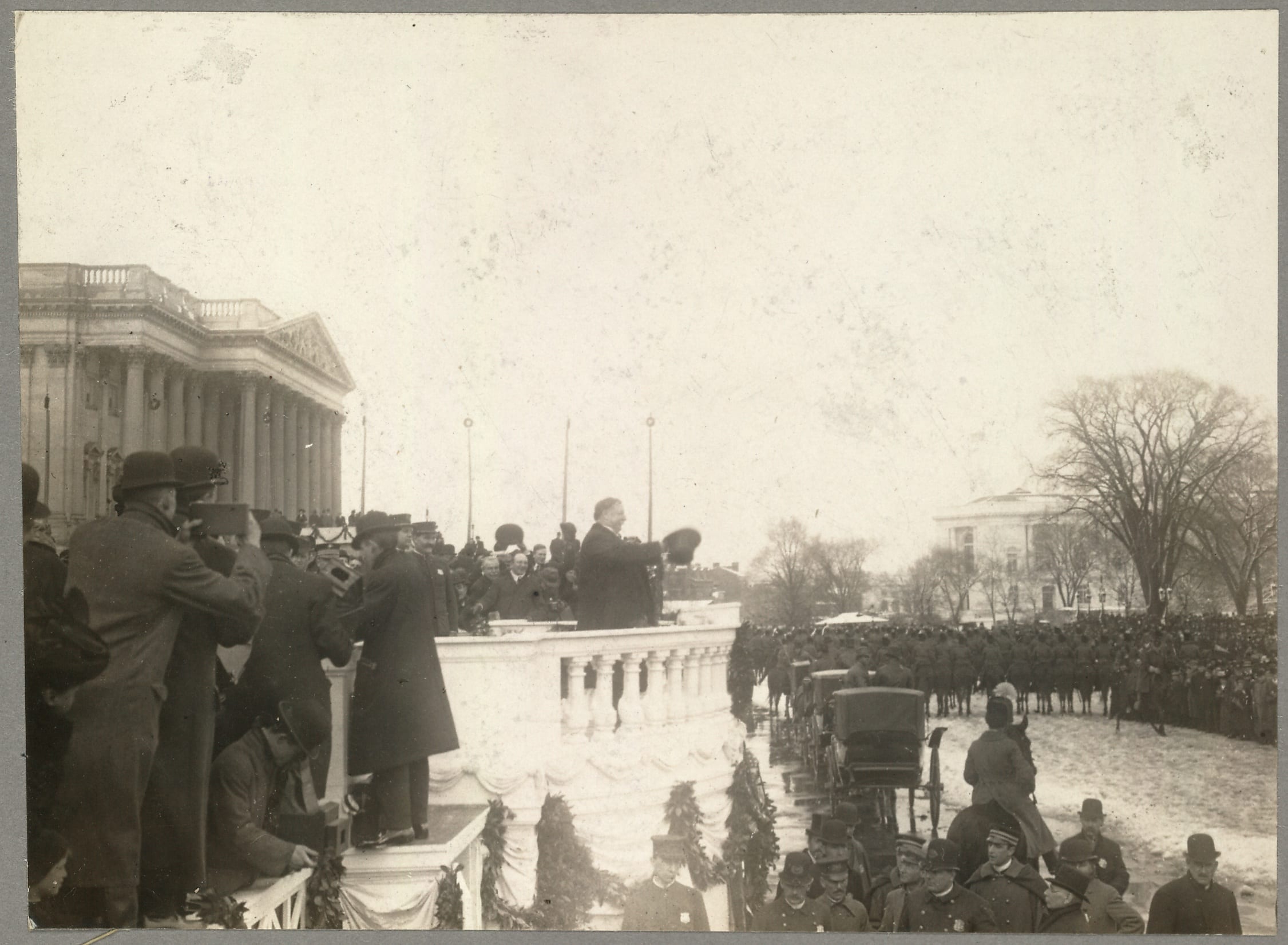 Side view of Taft on balustrade in snow, with top hat in hand, gesturing to crowd before him on grounds of east front of Capitol, with photographers at left.