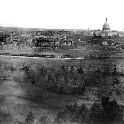 Looking toward the west side of the United States Capitol building, a panoramic view of the city of Washington, D.C. shows the Mall area in the foreground before the railroad tracks were removed. A train is visible on the tracks. The domed structure in front of the Capitol is the Botanic Garden original octagonal greenhouse of 1859 and behind it the central pavilion added after the Civil War. The buildings were razed in 1932. The Botanic Garden was on a site previously occupied by the Columbian Institute for the Promotion of Arts and Sciences in Washington, D.C.