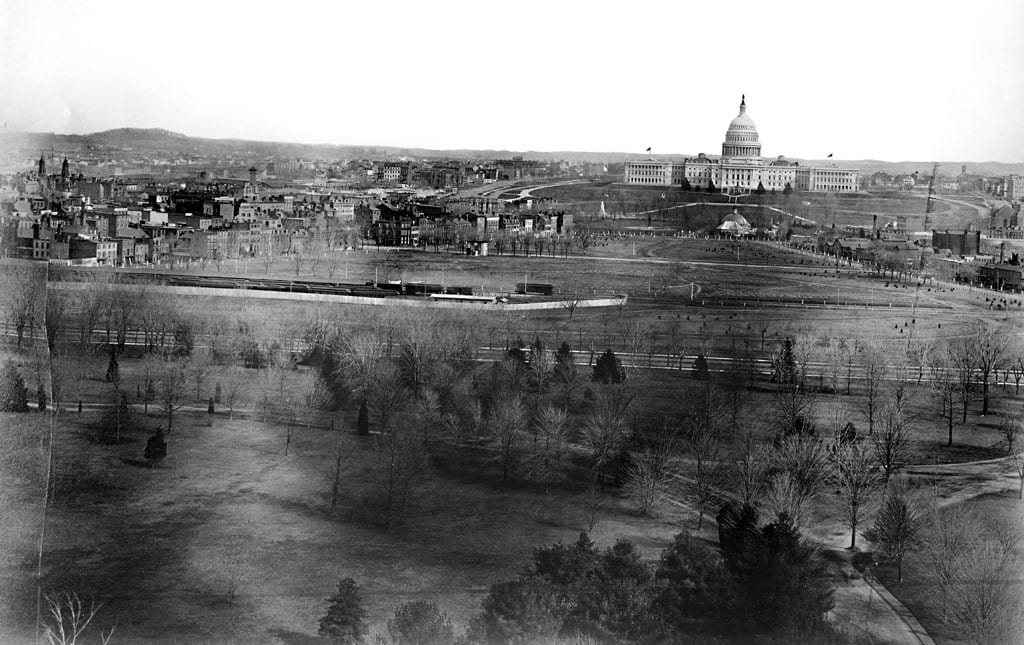 Looking toward the west side of the United States Capitol building, a panoramic view of the city of Washington, D.C. shows the Mall area in the foreground before the railroad tracks were removed. A train is visible on the tracks. The domed structure in front of the Capitol is the Botanic Garden original octagonal greenhouse of 1859 and behind it the central pavilion added after the Civil War. The buildings were razed in 1932. The Botanic Garden was on a site previously occupied by the Columbian Institute for the Promotion of Arts and Sciences in Washington, D.C.