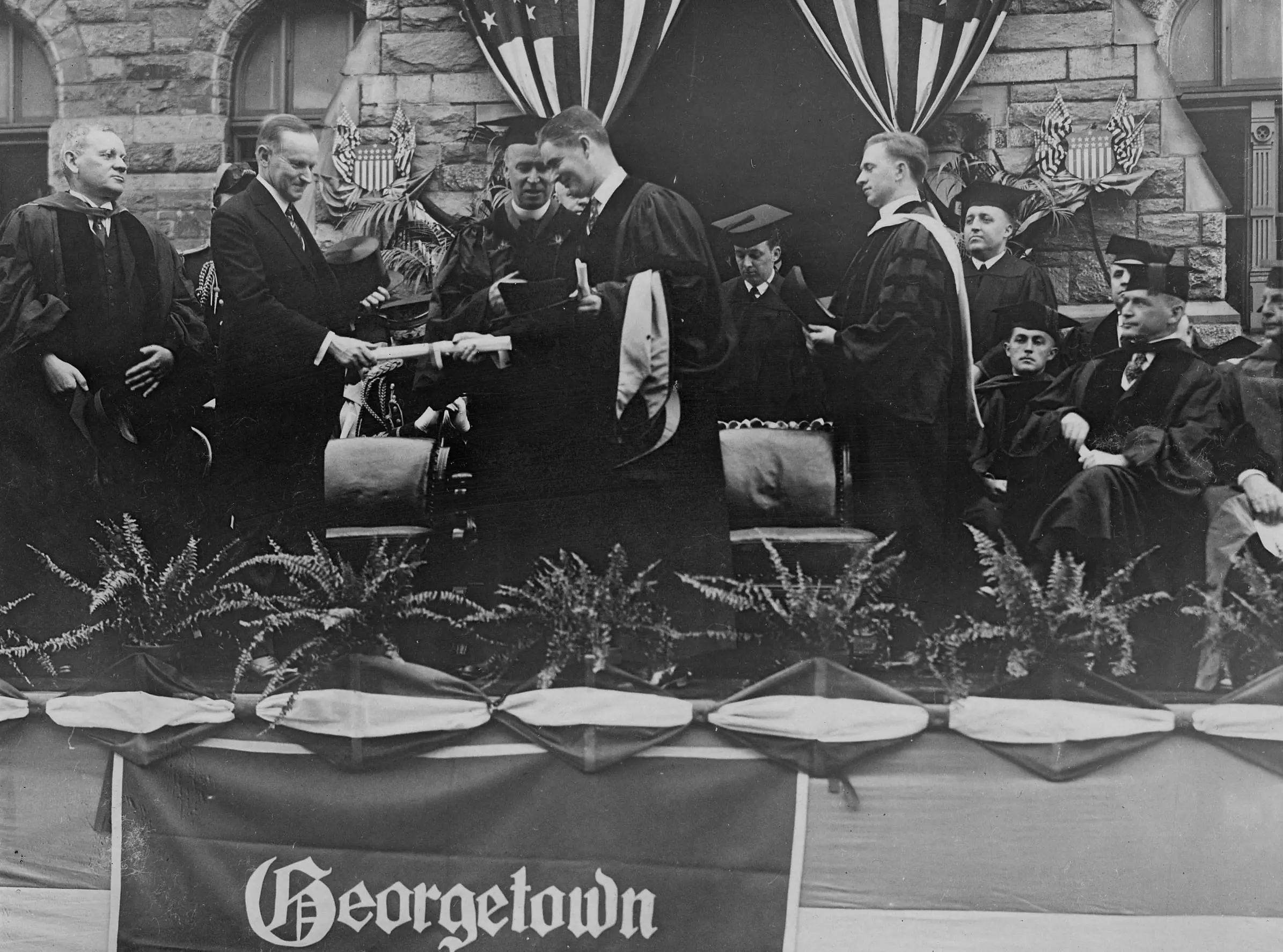 Calvin Coolidge, President of the United States, presents an honorary degree to Governor William S. Flynn of Rhode Island at the Georgetown University commencement. Pictured, left to right: Pierce Butler, Associate Justice of the Supreme Court; President Coolidge; John B. Creeden, S.J., President of Georgetown University; Governor Flynn; Walter J. O'Connor, University Registrar; and Andrew L. Bouwhuis, S.J.