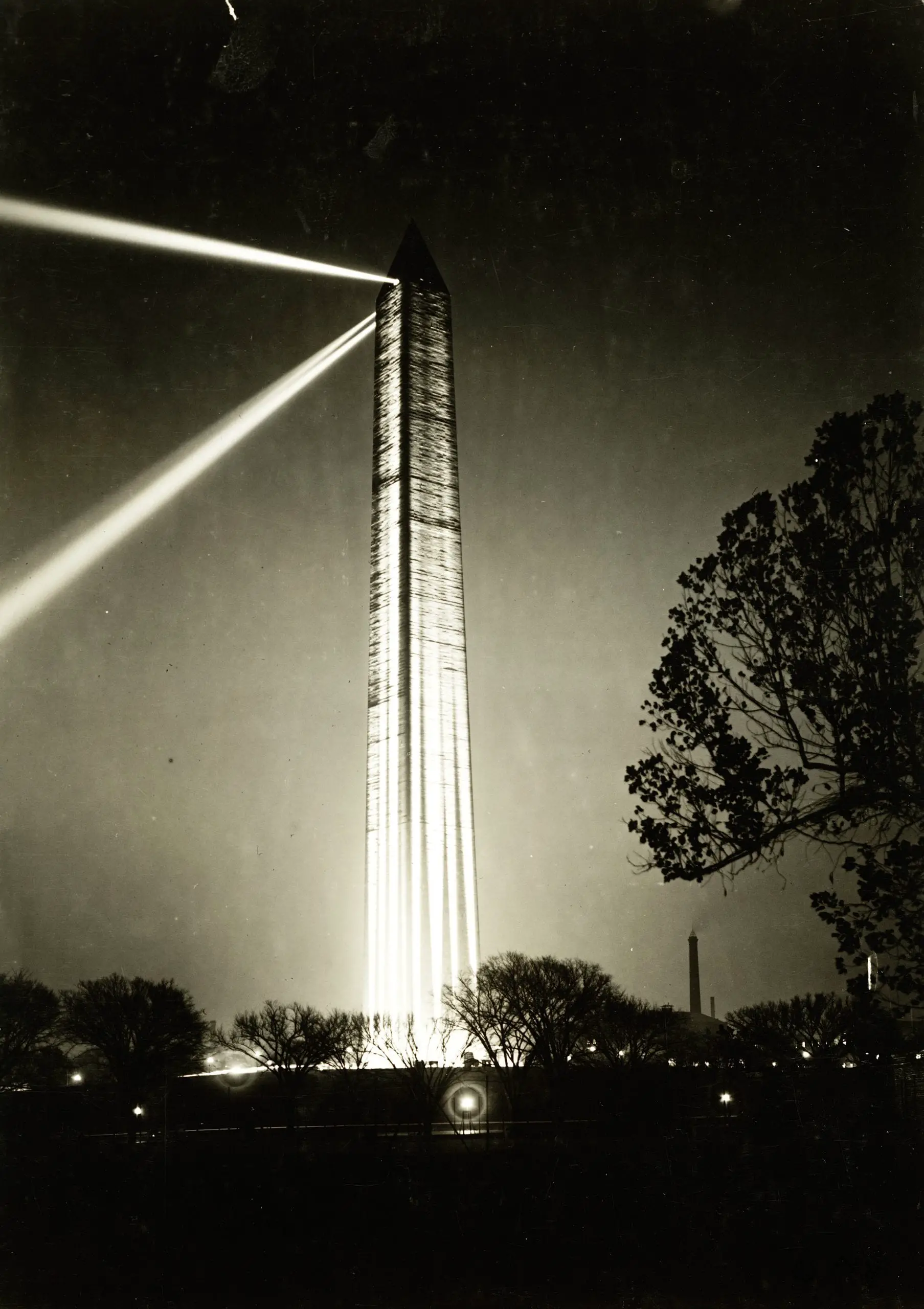 Photograph shows lights beaming from the top of the Washington Monument at night, also large lights on the ground, directed up, illuminating the sides.