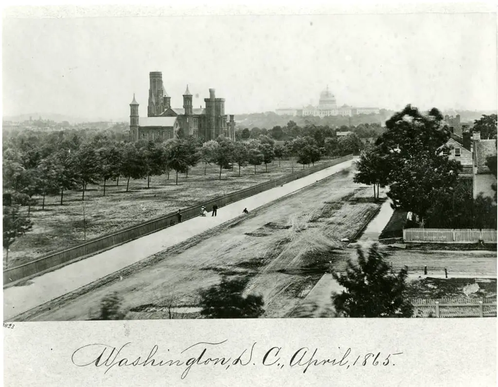 View of the Smithsonian Institution Building looking east up B Street (Independence Ave) towards the U.S. Capitol. Grounds are landscaped following a plan laid out by Andrew Jackson Downing, with the Castle nestled among trees. Visible amidst the trees is the Magnetic Observatory, built in 1853. People stand along the fence installed along the sidewalk on B Street In this image, the central roof of the Castle is intact and the small towers have the caps on them. In January of 1865, a fire destroyed the central roof and the caps on the towers, so this photograph had to have been taken prior to January 1865 and is most likely April 1863. The Capitol dome, seen in the distance, is not yet completed