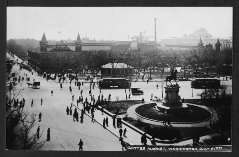 Expansive view of Center Market taken from an elevated position on the northeast corner of Indiana Avenue and 7th Street NW. A statue of Major General Winfield Scott Hancock is prominnent on the right side of the image which also features pedestrians, streetcars, horse and carriages, bicycles, and automobile traffic.