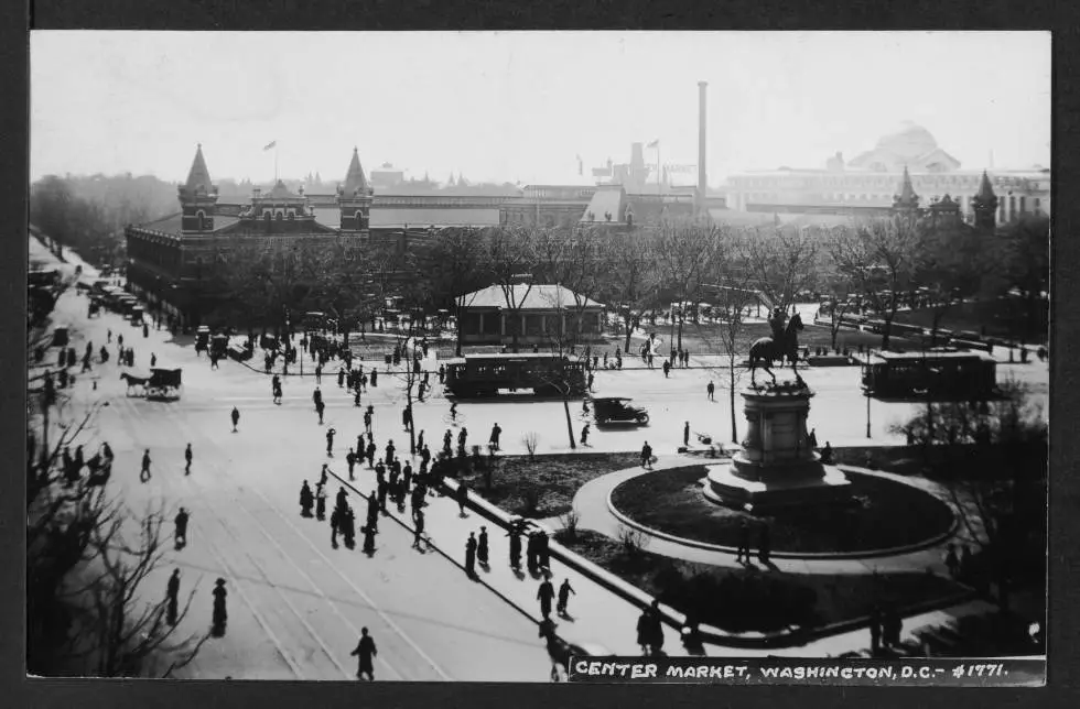 Expansive view of Center Market taken from an elevated position on the northeast corner of Indiana Avenue and 7th Street NW. A statue of Major General Winfield Scott Hancock is prominnent on the right side of the image which also features pedestrians, streetcars, horse and carriages, bicycles, and automobile traffic.