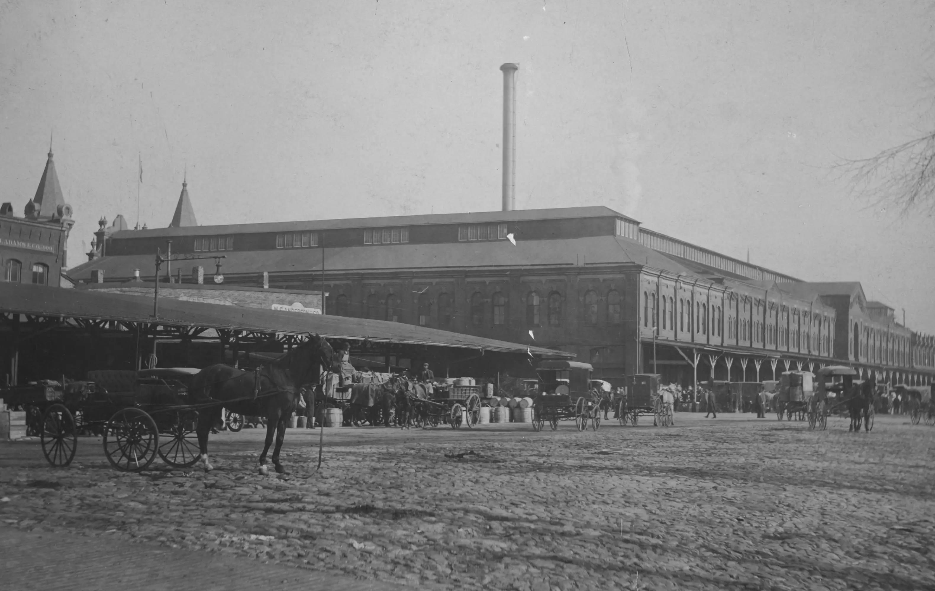 View of Constitution Avenue NW looking towards Center Market, which occupied the northeast corner of 9th Street NW. Many produce stalls are visible on the northwest corner of that intersection and horse-drawn vehicles are on the cobblestone street.