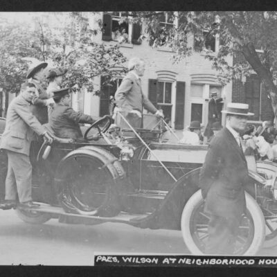 Woodrow Wilson stands in the front of a convertible automobile stopped in the intersection of what was then N and Union Streets SW. Wilson, the driver of the car, and two woman in the back look east at something happening on N Street. A crowd gathered under a tree near the car and people in front of 485 N Street also look in the same direction. Two Secret Service agents look at the photographer. Written on the reverse is "Barney Neighborhood House, 470 N Street SW." Wilson had only been President of the US for two months when the photo was taken.
