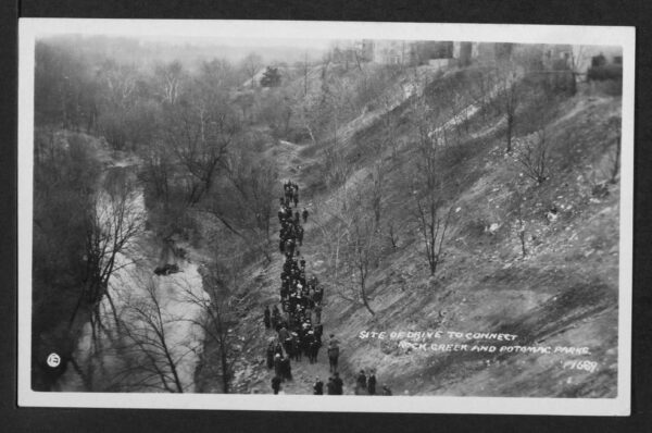 View of a group of hikers walking alongside Rock Creek to the left and an embankment that leads up to the right upon which can be seen a row of buildings. Location is to the west and downhill from the 2800 block of Adams Mill Road NW.