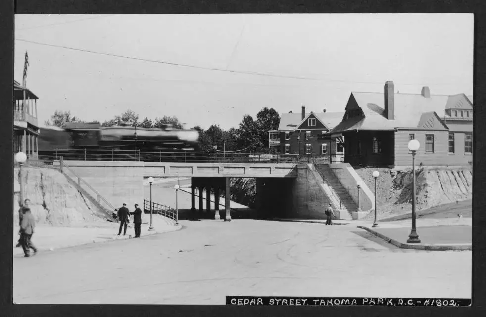 View looking from the northwest corner of Cedar and 4th Street NW east down Cedar Street toward the railroad underpass. Image includes a partial view of the Baltimore & Ohio Railroad station on the right with a blurred locomotive crossing the underpass and going into the station. The train is headed toward Washington, D.C.