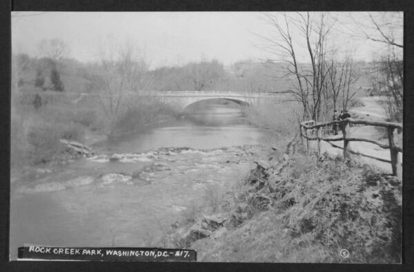 Wide view of the Old Harvard Street Bridge across Rock Creek Park taken from the south (down creek) side. A dirt road with a log fence appears on the right.