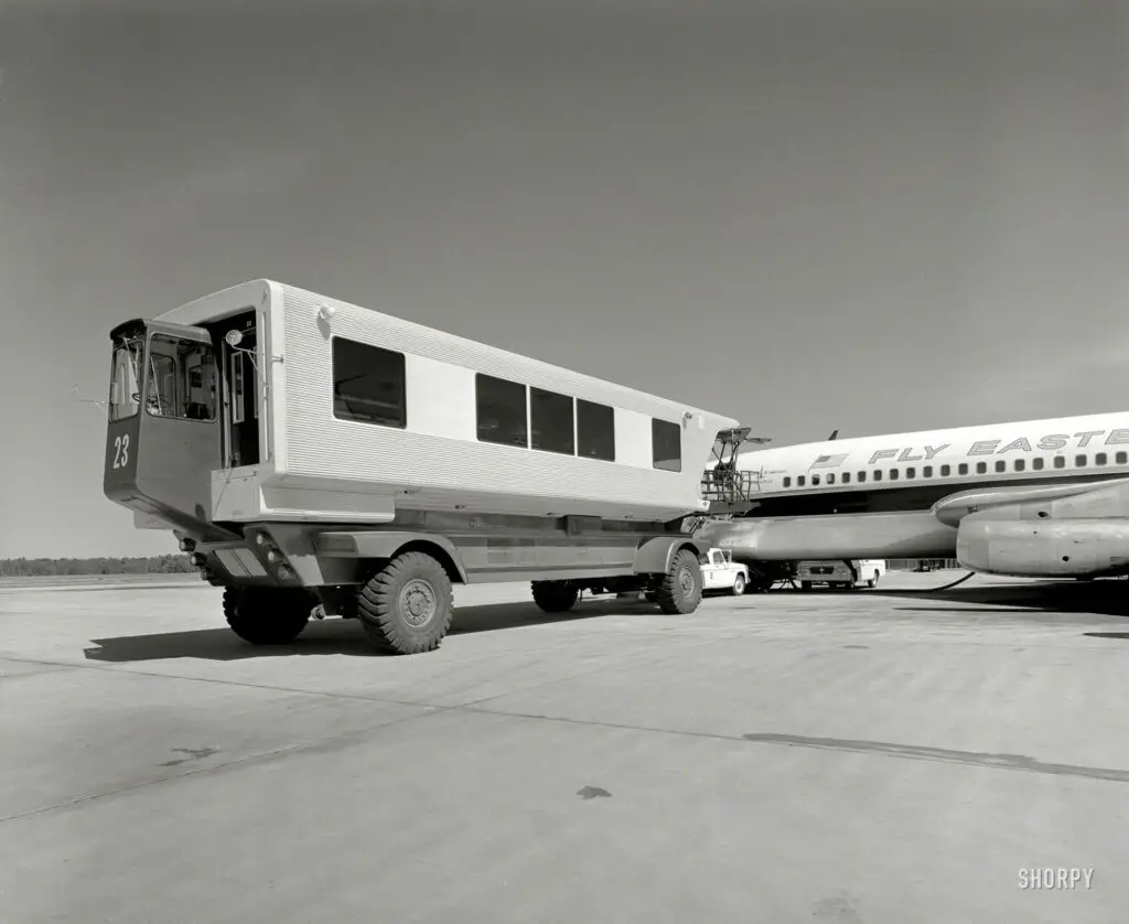 Black and white photograph depicting a mobile lounge vehicle with the number '23' prominently displayed, positioned next to an aircraft with the words 'Fly Eastern' on its body, on the tarmac at Dulles Airport shortly after it was built.