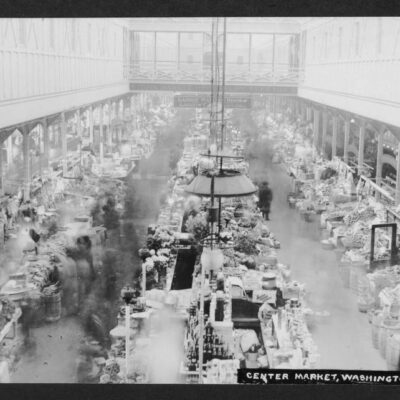 Interior view of Center Market showing the interior architecture of the market, a variety of stalls, blurred people, and signs reading "THIS MARKET OPEN EVERY WEEK DAY" and "LADIES' WAITING ROOM LADIES'... 7th St. WING TAKE ELEVATOR."