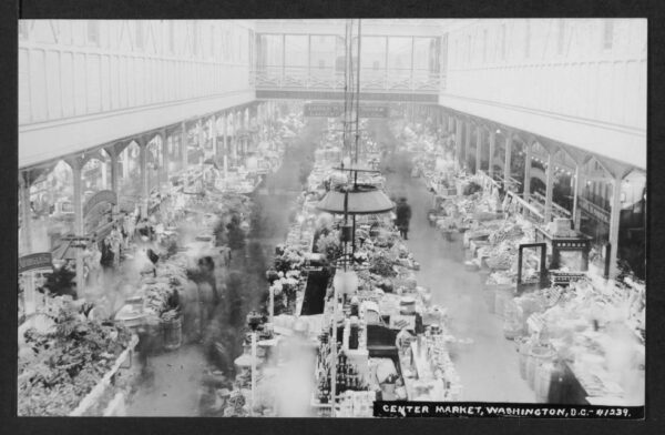 Interior view of Center Market showing the interior architecture of the market, a variety of stalls, blurred people, and signs reading "THIS MARKET OPEN EVERY WEEK DAY" and "LADIES' WAITING ROOM LADIES'... 7th St. WING TAKE ELEVATOR."