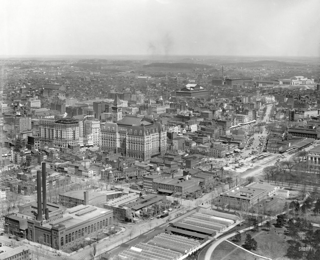 Circa 1911. "Washington from Washington Monument." Points of interest in this panoramic view include B Street (today's Constitution Avenue), running diagonally from the Potomac Electric powerhouse at lower left; Louisiana Avenue, branching off in the general direction of Union Station at upper right; the Old Post Office and its clock tower at left-center across Pennsylvania Avenue from the Raleigh Hotel under construction; the Agriculture Department greenhouses in the foreground with a corner of the Smithsonian "National Museum" at far right, just below Center Market; Liberty Market at upper left, below what looks to be a vast tent encampment; and, at right-upper-center, the Pension Office north of Judiciary Square and the District Court House. 8x10 glass negative, Detroit Publishing Co.