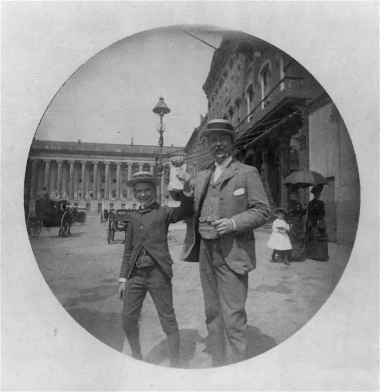 Photo shows a man and a boy in summer straw hats happily displaying some unidentifiable papers on F Street, with the Treasury and a new electric hooded arc streetlight in the background. One of a series of scenes near Painter's office at 14th and E streets, this picture bears the hallmarks of the new "snapshot" style now made possible by the invention of the quick, hand-held Kodak: natural smiles and the movement of the ladies under the umbrella.
