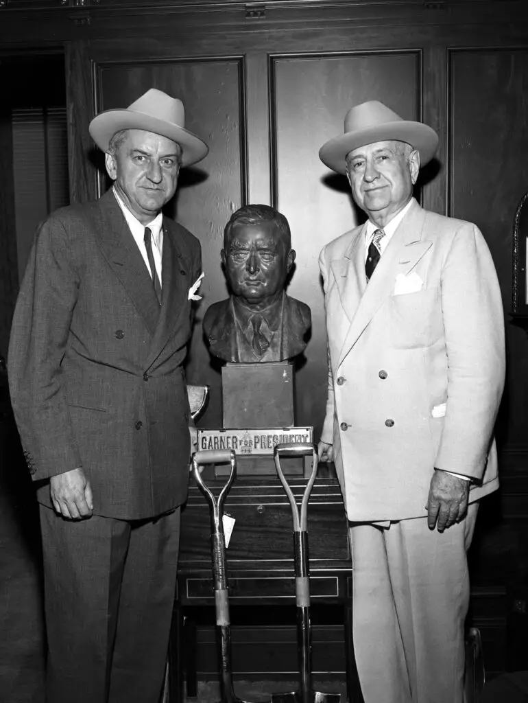 George Preston Marshall (left) and Amon Carter standing next to a bust of John Nance Garner. There are two shovels at the bottom of the bust pedestal. Both men are dressed in suits, neckties and hats. Mr. Marshall is the owner of the Washington Redskins.