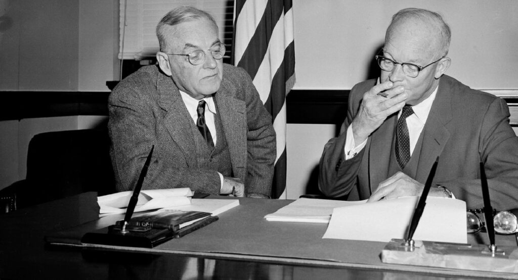 Black and white photo of John Foster Dulles and Dwight D. Eisenhower seated at a desk, engaged in a discussion.