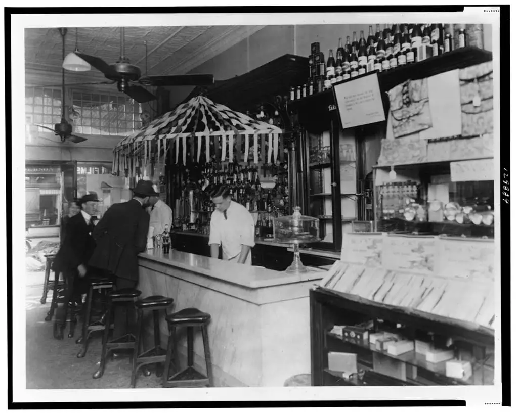 Vintage black-and-white photograph of People's Drug Store's interior at 11th and G Streets, Washington, D.C. Employees are seen standing behind the counter of a soda fountain, attending to seated customers.
