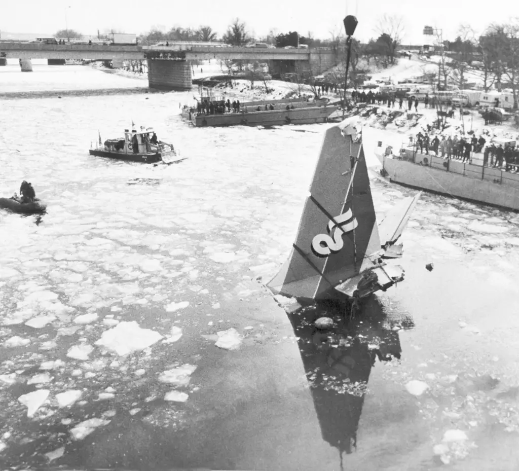 Work crews use a tall crane to raise the destroyed tail piece of an airplane out of a river, with a bridge seen damaged and covered in snow in the background where the aircraft initially hit.