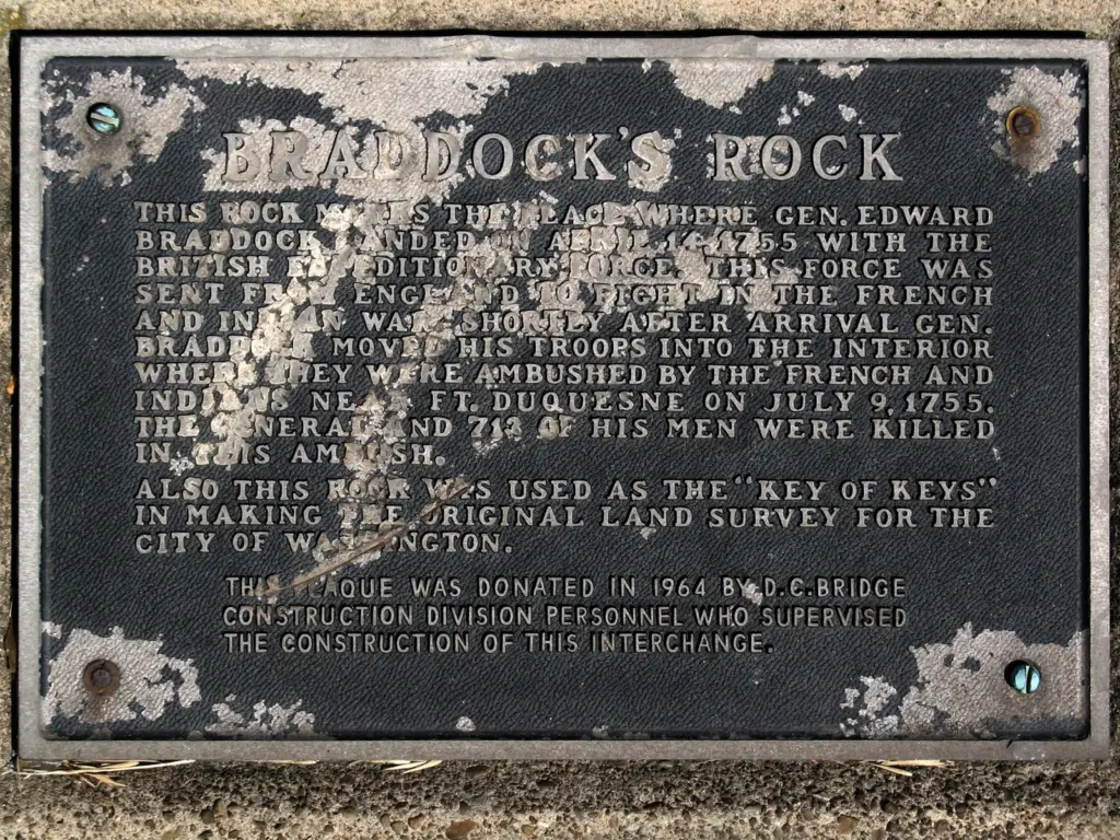 A metal plaque mounted on a stone base, engraved with aged text commemorating Braddock's Rock. The plaque sits overgrown with weeds and vines beside a highway bridge support pillar. Its worn lettering reads: "Here at Braddock's Rock, adjacent to this plaque, landed British and Colonial troops in April 1755 en route to drive the French from Fort Duquesne in the first engagements of the French and Indian War." Below is a dedication from D.C. Bridge Construction Division personnel who directed infrastructure upgrades impacting the rock remnants in that vicinity.