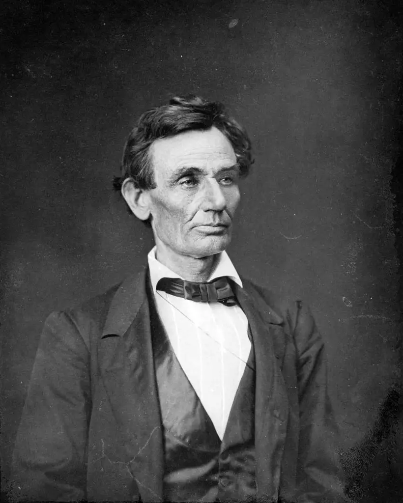 Black and white photograph of Abraham Lincoln from 1860. Lincoln is shown from the chest up, wearing a dark suit with a white shirt and black bow tie. His face is solemn, with deep-set eyes and visible lines, characteristic of his later portraits.