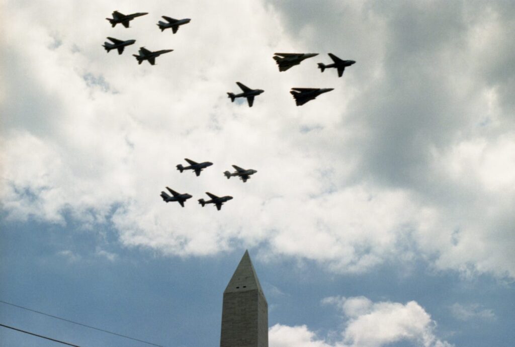 A formation of military aircraft soars overhead the Washington Monument during the National Victory Celebration parade on June 8, 1991. The jets, representing planes used in Operation Desert Storm, trail contrails against a clear sky as spectators watch from below.