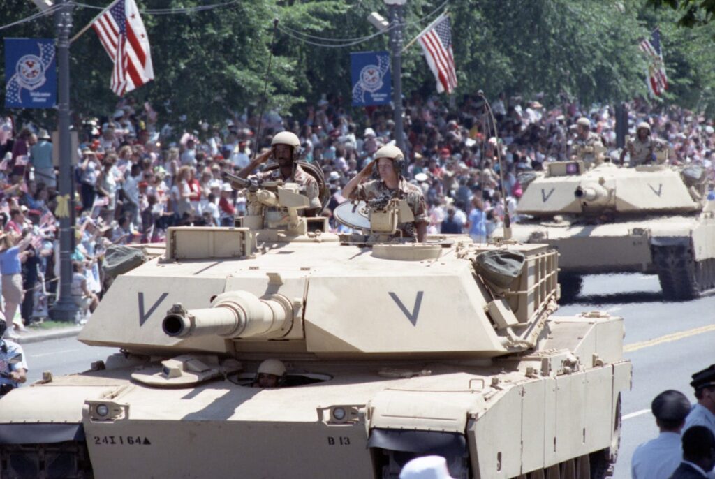 A line of M1A1 Abrams tanks rolls down Constitution Avenue during the National Victory Celebration parade in Washington, D.C., June 8, 1991. The tanks are combat-loaded and meticulously aligned, flanked by cheering spectators waving American flags along the parade route.