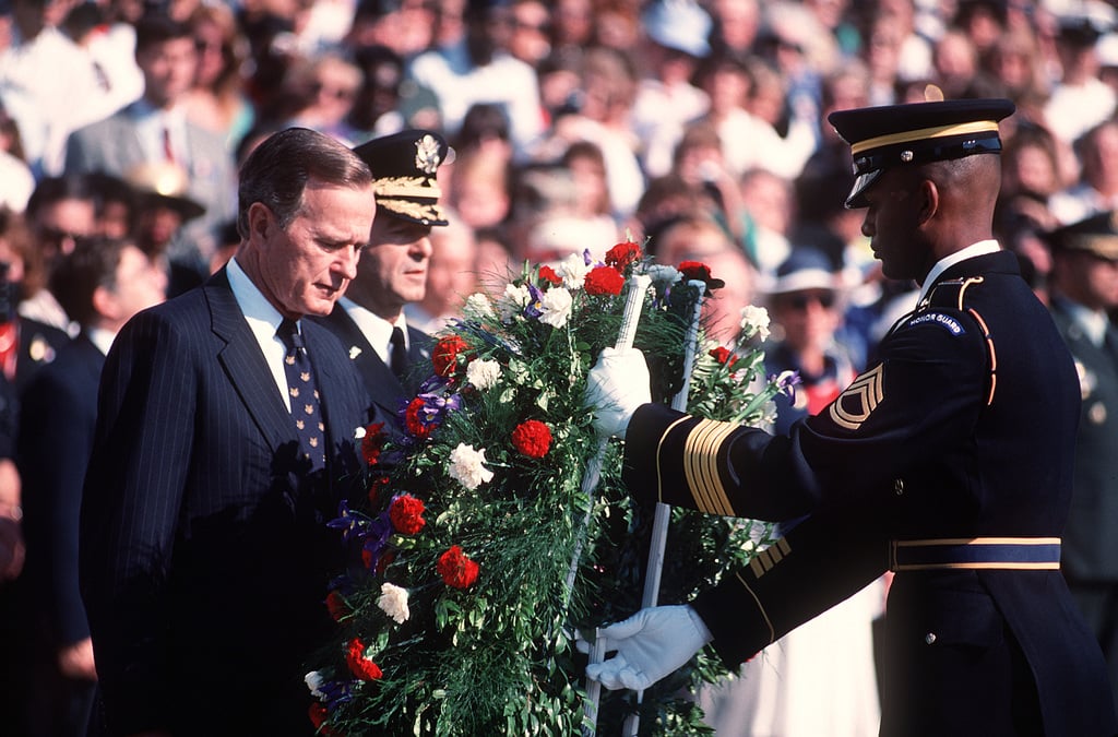 President George H.W. Bush, dressed in a dark suit, places a funeral wreath on a stand before the Tomb of the Unknown Soldier at Arlington National Cemetery. A solemn military honor guard stands at attention in the background, framed by ceremonial flags and evergreen trees.
