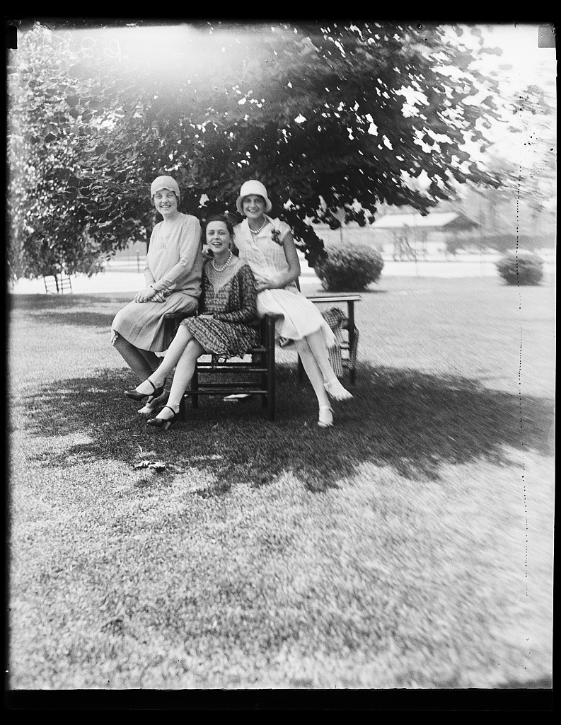 Three women in 1928 dresses sitting on a garden bench under a tree at the Chevy Chase Club