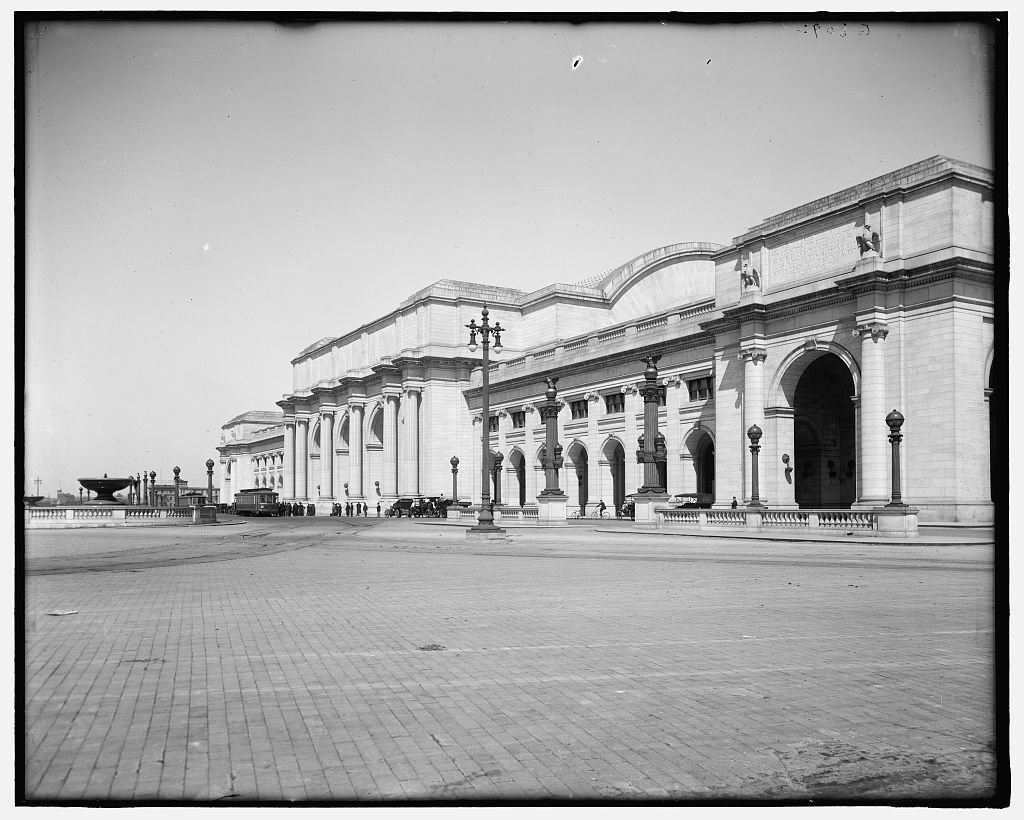 Front facade of the newly-completed Union Station in Washington DC shortly after its 1908 opening, a white Beaux-Arts station with columns and arched entrances, with a streetcar and early automobiles in the plaza