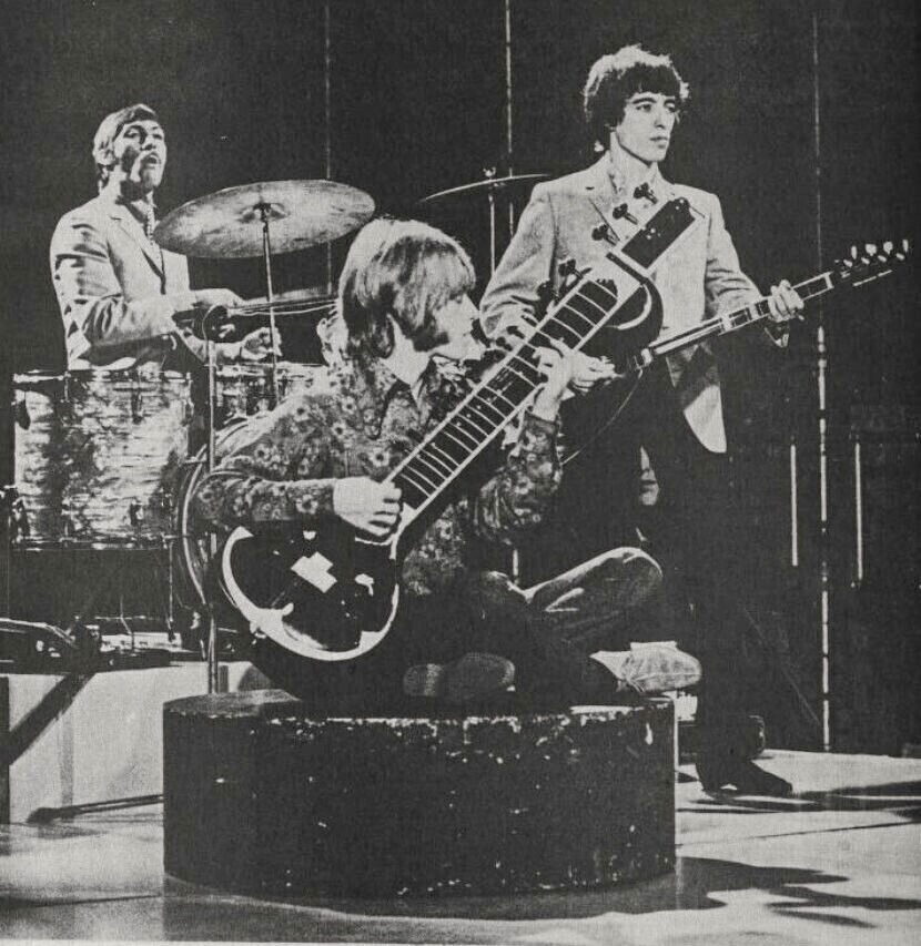 Brian Jones of the Rolling Stones sitting cross-legged on a small round stage playing a sitar, with Charlie Watts on drums behind him.