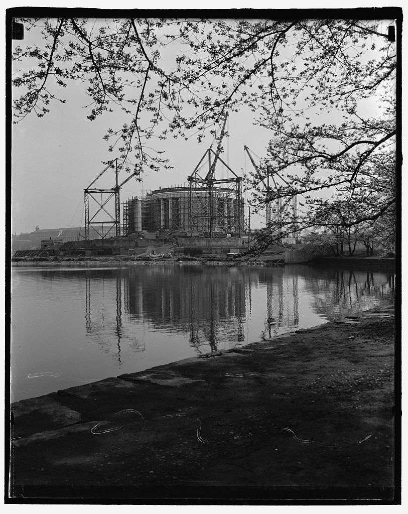 The Jefferson Memorial under construction across the Tidal Basin, with cranes around the unfinished dome, framed by cherry blossom branches.