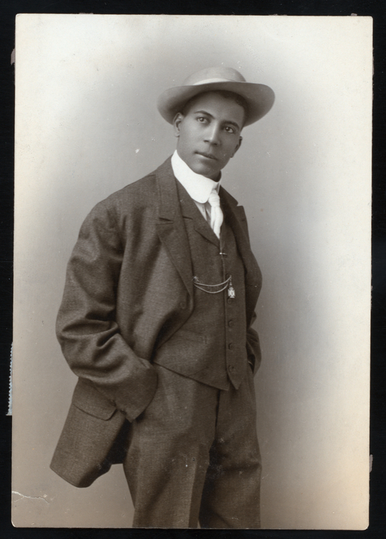 Black-and-white studio portrait of Sherman H. Dudley, a young Black man in a three-piece suit and wide-brimmed hat with a watch chain, standing with one hand in his pocket.