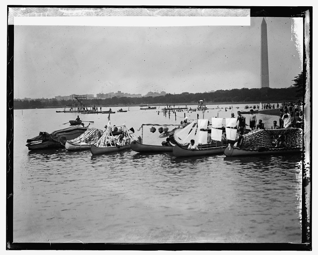 Decorated canoes float on the Tidal Basin in front of the diving platform, with the Washington Monument rising in the distance and crowds of swimmers along the shore.