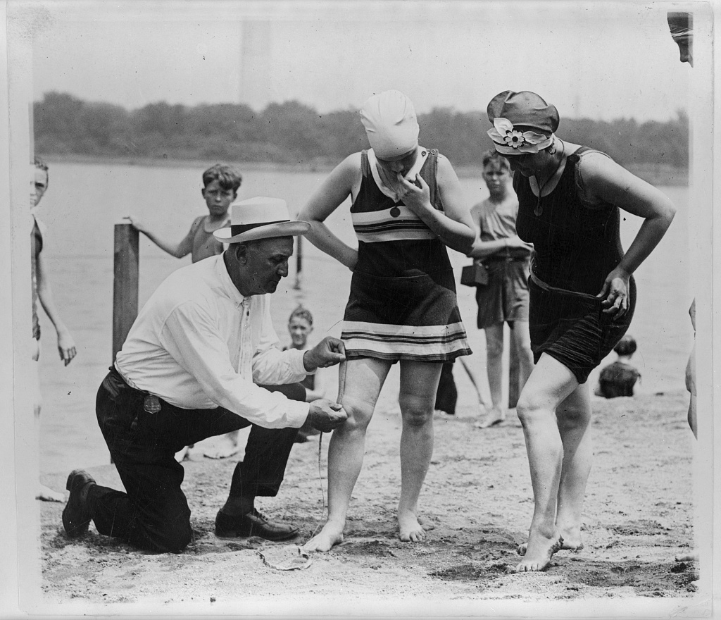 A police officer kneels on the sand to measure the bathing suit length on two women at the Tidal Basin beach, with the Washington Monument in the background.