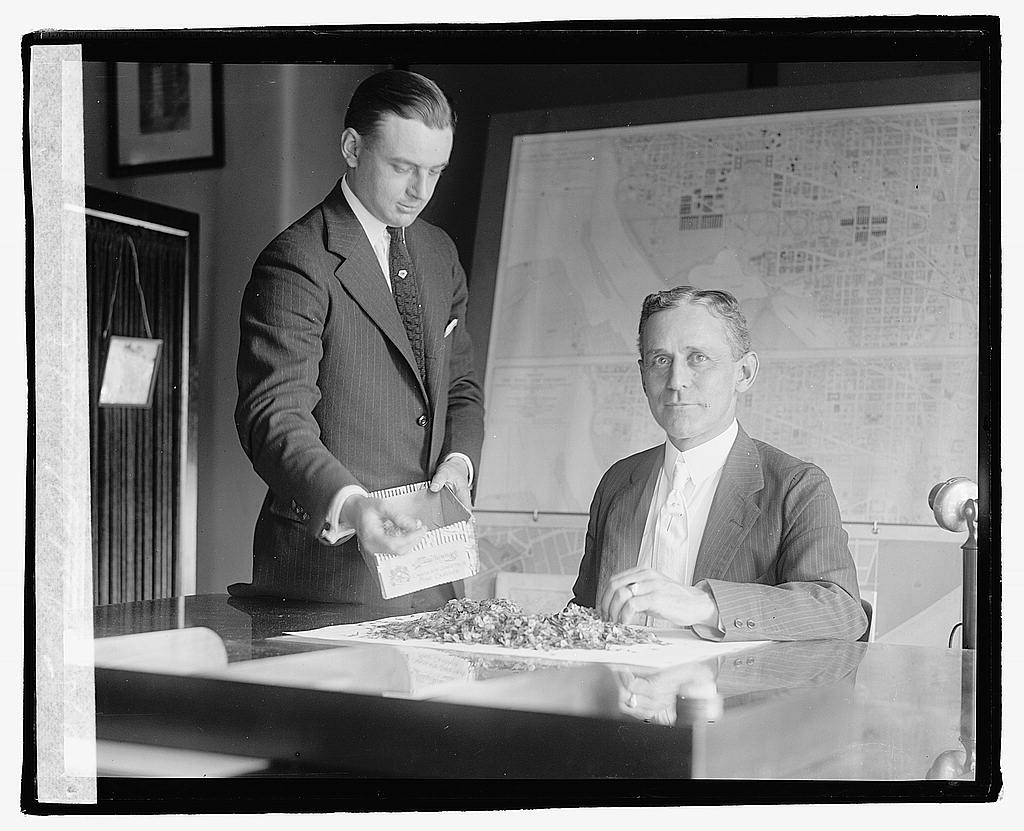 Colonel Clarence O. Sherrill sits at a desk in a Washington office. A large detailed map of the District of Columbia covers the wall behind him. A second man in a suit stands beside the desk.