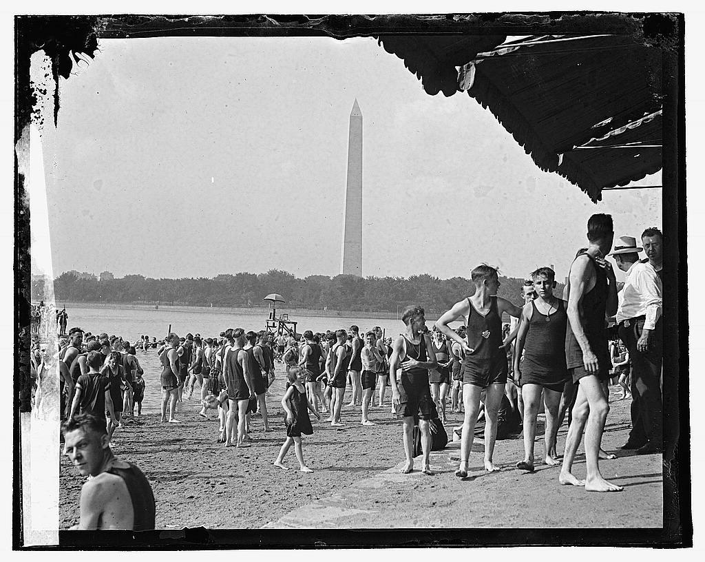 A wide view of swimmers along the sandy bank of the Tidal Basin with the Washington Monument rising in the distance.