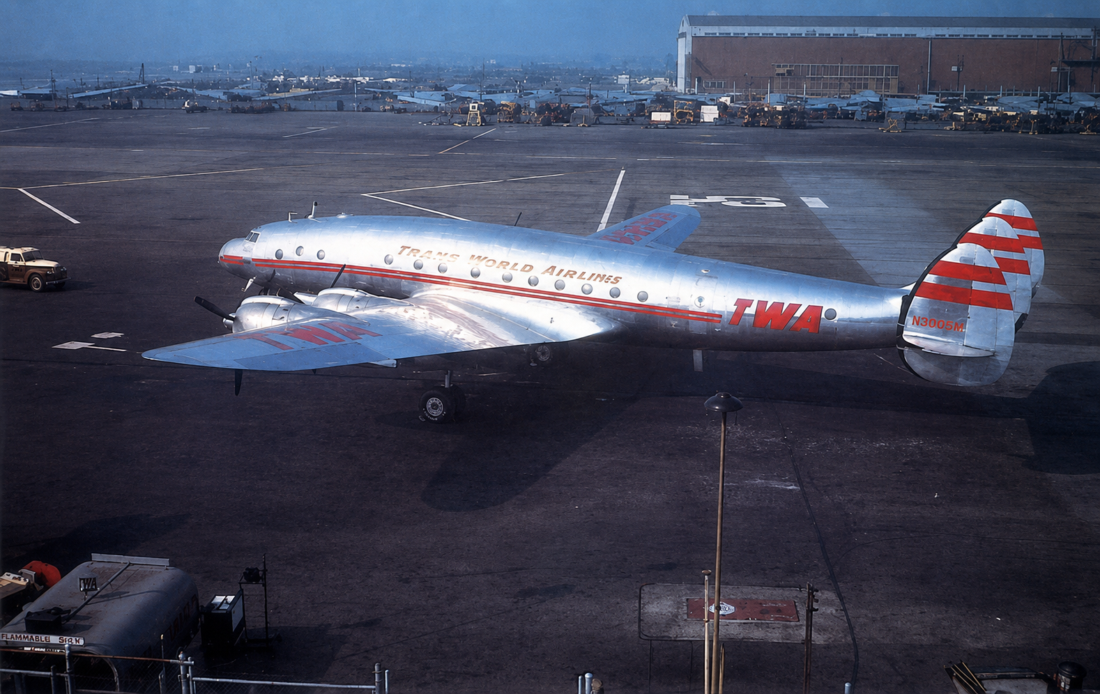 Color photograph of a Trans World Airlines Lockheed L-049 Constellation named Paris Sky Chief (NC86505) parked on an airport ramp. The polished silver aircraft displays TWA markings and the distinctive triple-tail design, with hangars and airport equipment visible in the background.