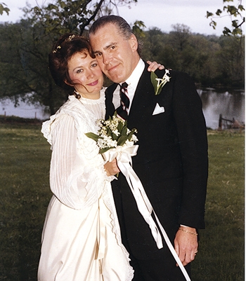 Carol and Howard Joynt on their wedding day, with Carol in a white gown holding flowers and Howard in a black tuxedo, photographed outdoors