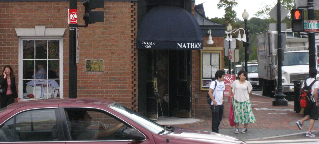 Daytime exterior of Nathans bar at the corner of Wisconsin Avenue and M Street NW in Georgetown, with the dark blue NATHANS awning, The Q and A Cafe subtitle, and pedestrians on the brick sidewalk