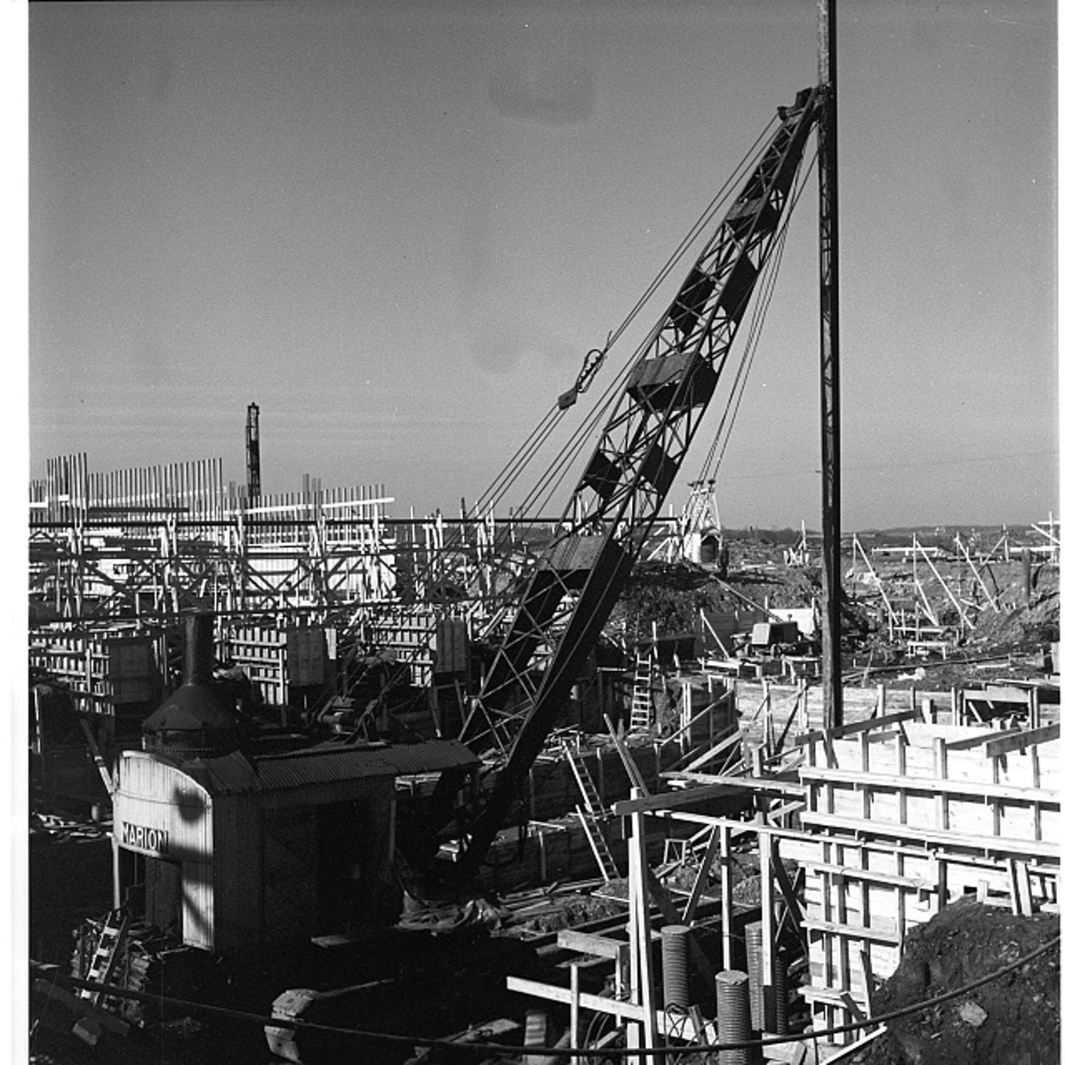 A Marion crane lifts steel forms over the rising concrete walls of the Pentagon, with cars and rising support towers visible in the distance, 1942