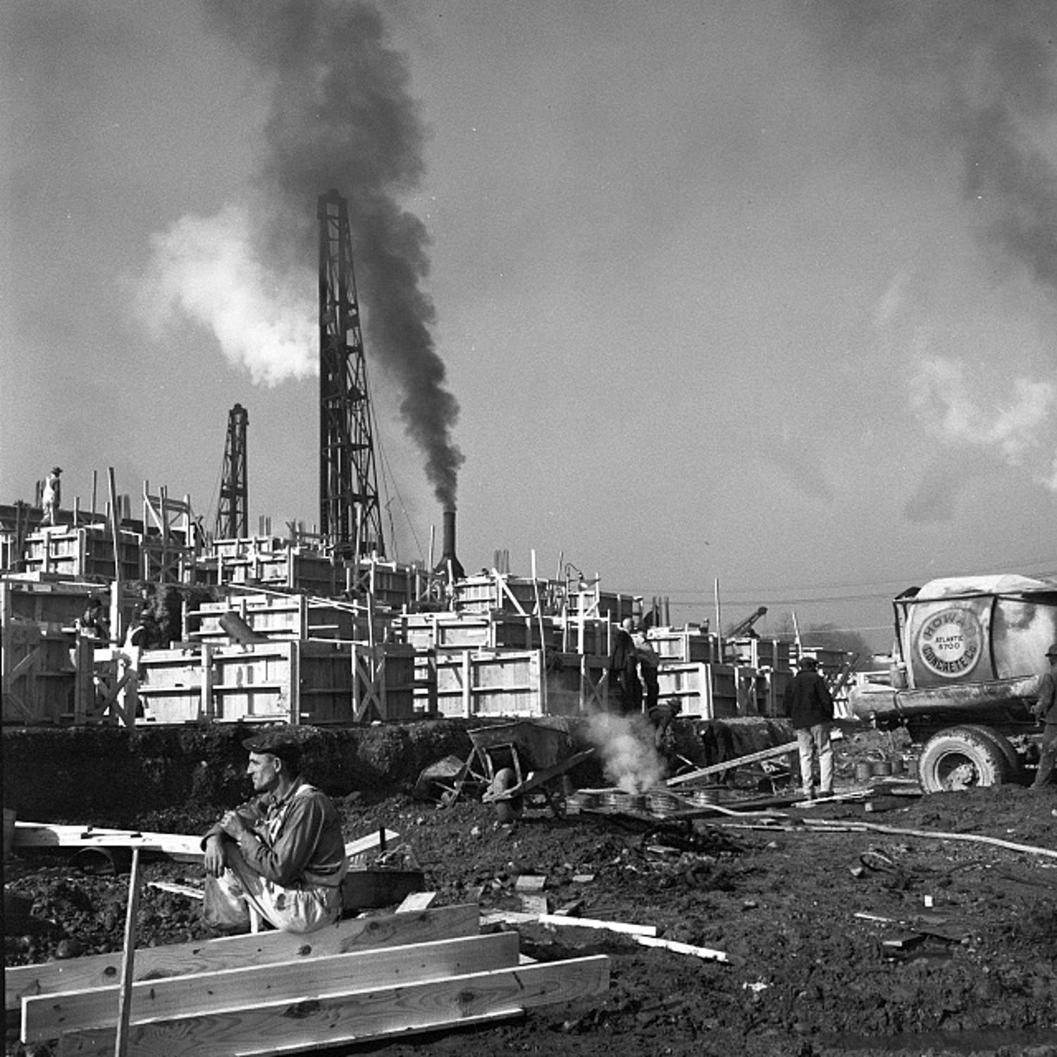 Construction worker rests on lumber at the Pentagon site as smokestacks and concrete forms rise behind him, 1942