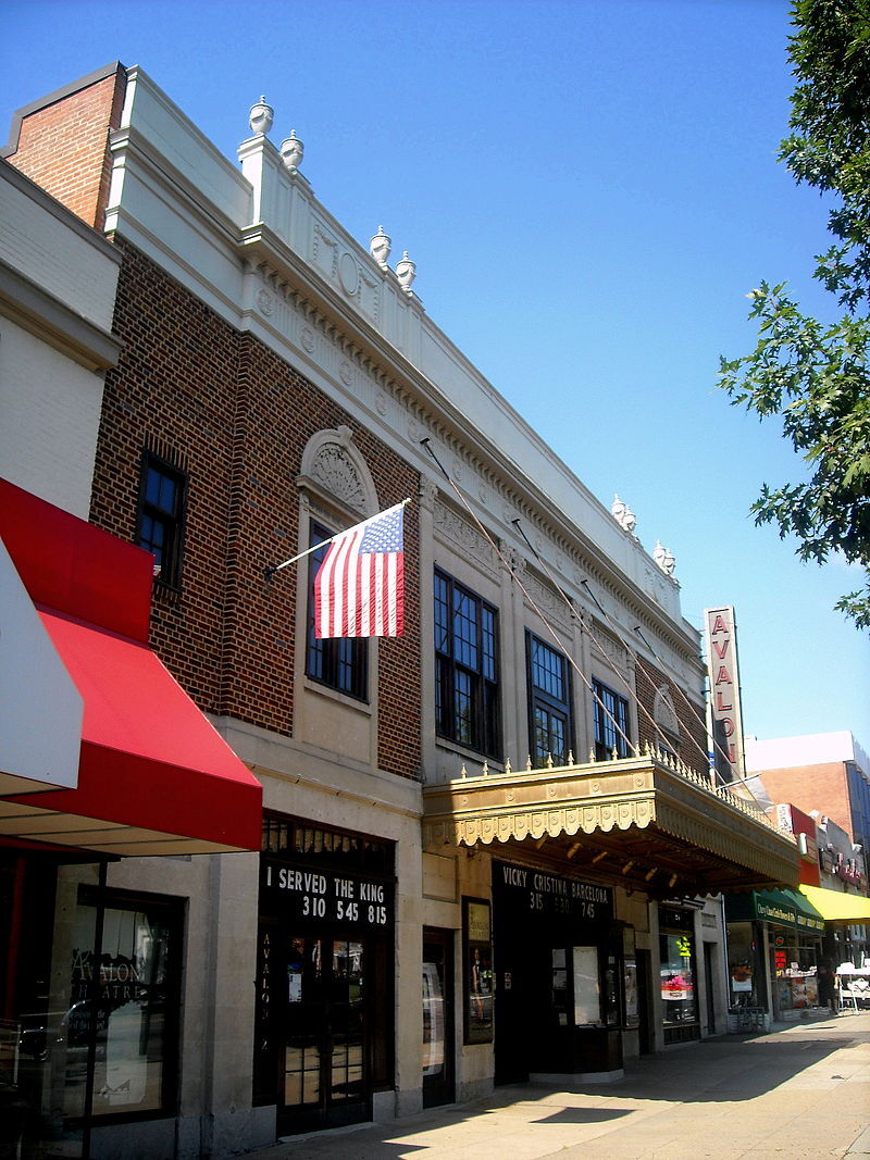Angled view of the restored Avalon Theatre facade with vertical Avalon sign, brick and limestone detailing, an American flag, and a marquee showing Vicky Cristina Barcelona.