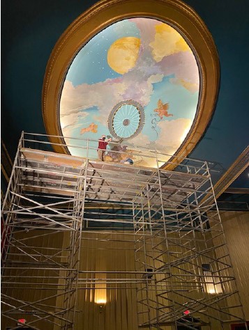 Circular ceiling mural of Mercury tossing a film reel to a cherub, seen from below with restoration scaffolding in the foreground.