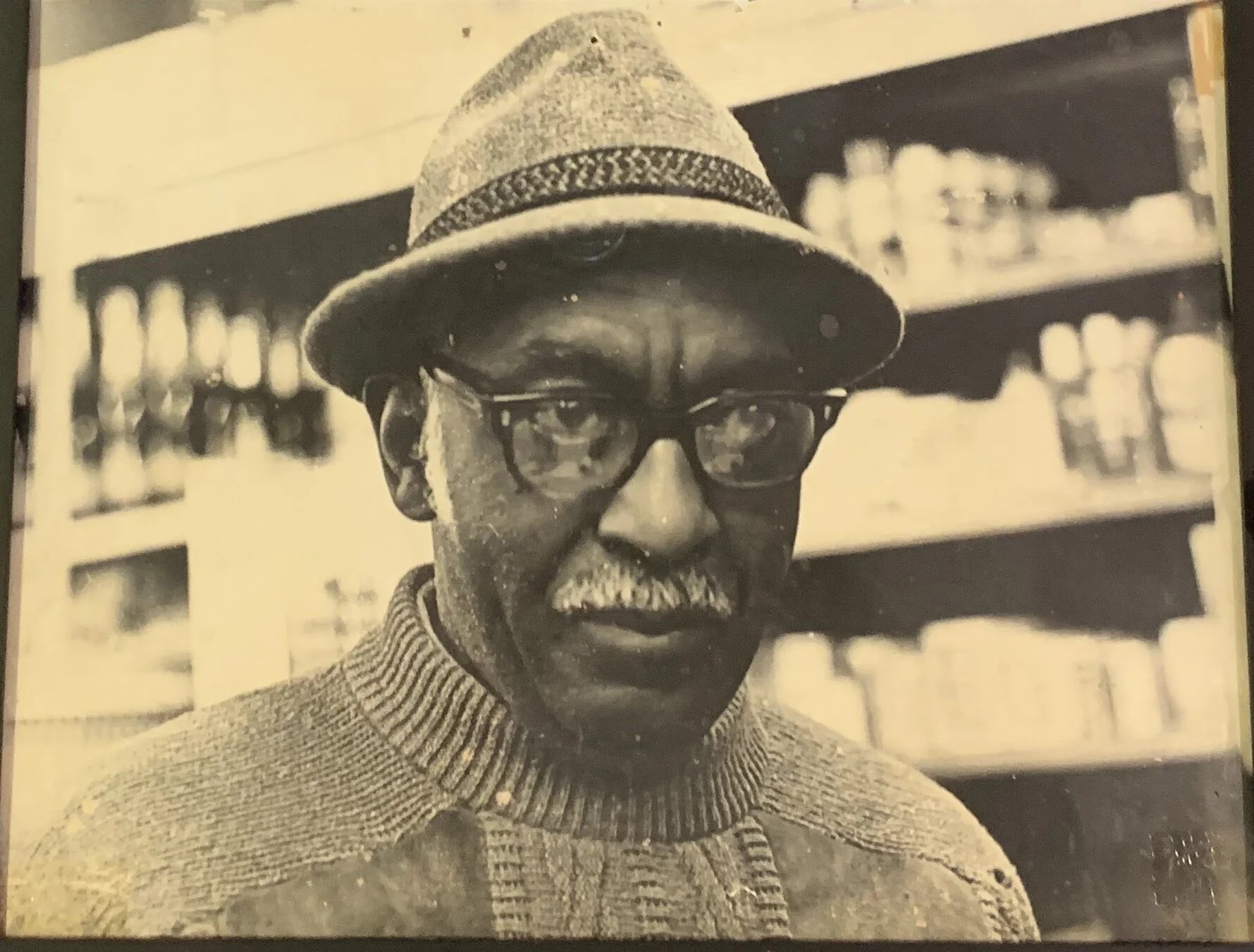 Sepia toned close portrait of an older Black man wearing a tweed fedora, glasses, and a knit sweater, photographed in front of the market's grocery shelves.