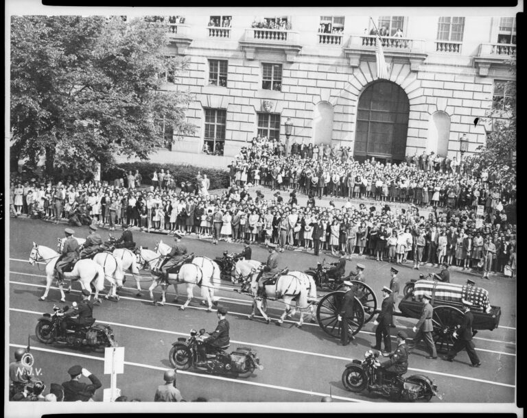 A horse-drawn caisson carries a flag-draped coffin past crowds lining the street.